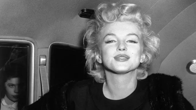 Una Marilyn Monroe sonriente mira a cámara. A la izquierda se vislumbra otra persona,.