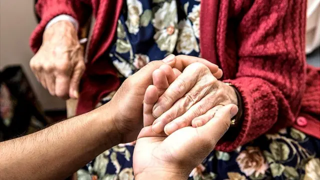 የዕድሜ ባለጸጋን ሰው እጅ የያዘች የእንክብካቤ ሰጭ ሰራተኛ 