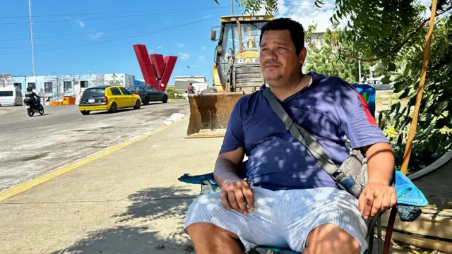 Enyerber Pernalete, de 44 años, cerca de su torre. Lleva una chemise azul y un short azul con rayas blancas. Al fondo se ve una gran V, símbolo de la Misión Vivienda. 