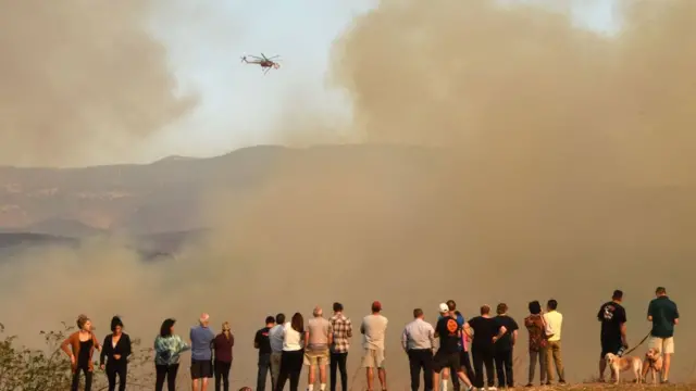Crowd of people dey look from far how fire-fighters dey try off di fire from helicopter.