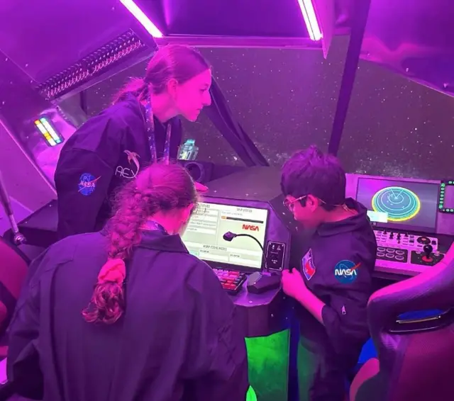 David Camacho, con otras dos mujeres jóvenes, todos vestidos con overoles de la NASA, están dentro de una cabina especial observando una computadora