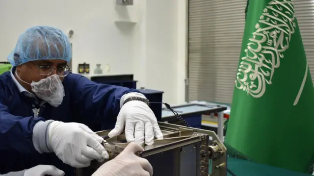 صورة نشرتها وكالة الفضاء السعودية لما قالت إنهم مهندسون سعوديون يرتدون قفازات وأقنعة وجه وأغطية رأس يعملون على القمر الصناعي شمس بجوار علم المملكة الأخضر