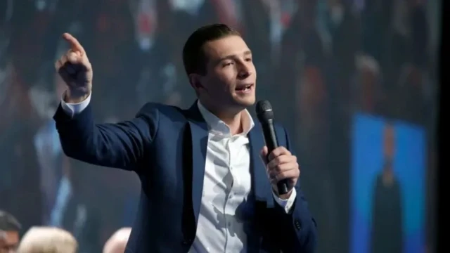 Молодой мужчина в расстегнутом пиджаке и белой рубашке с расстегнутым воротником, подняв правую руку, что-то говорит в микрофон