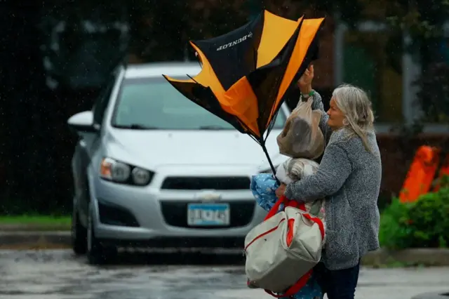 Mulherpoker aestacionamento segurando sacolas e guarda-chuva quebrado por conta dos ventos