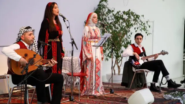 خلود إبراهيم، ترتدي لباساً تقليدياً، وتغني مع كورال "ناي" في حفلة موسيقية إلى جانب عازفين.