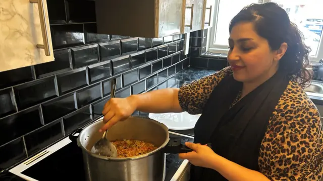 Nelab preparing a meal in her kitchen in a large pot with a ladle. She is wearing a leopard-print top and black scarf.