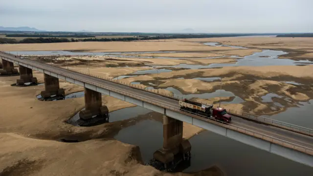 A imagem mostra um caminhão passando por uma grande ponte sobre um enorme banco de areia com pequenas ilhas de água, fruto da seca