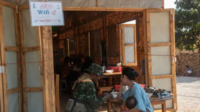 စစ်တပ်ကို ဆန့်ကျင်တိုက်ခိုက်နေသည့် လက်နက်ကိုင်အဖွဲ့များ ထိန်းချုပ်ရာ ကယား (ကရင်နီ)ဒေသတွင် Starlink အင်တာနက် အသုံးပြုနိုင်ကြောင်း ဖော်ပြထားသည့် ဆိုင်းဘုတ်အနီး လမ်းဘေးစားသောက်ဆိုင်တစ်ခု၌ တော်လှန်ရေးတပ်ဖွဲ့ဝင်တစ်ဦးနှင့် မိသားစုကို တွေ့ရစဉ်