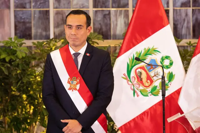 José Jeri, presidente de Perú, durante la ceremonia de juramento de sus nuevos ministros en el Palacio de Gobierno en Lima, vestido de traje de chaqueta, manos cruzadas por delante y la banda presidencial con los colores de Perú, rojo y blanco, cruzada en el pecho. A su lado hay un micrófono de pie y, detrás, dos banderas grandes de Perú. 