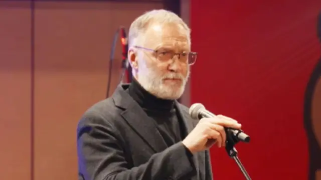 Rus adam gri bir takım elbise ve siyah balıkçı yaka kazak giymiş, gözlük takmış, kısa gri saçlı ve kısa kesilmiş gri sakallı. Sahnede duruyor ve elinde bir mikrofon tutuyor.