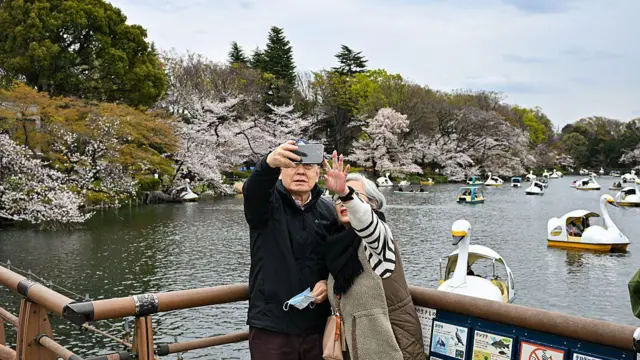 Um casal de idosos tira uma selfie com o celular em frente a um lago cheio de barcos e flores de cerejeira no Parque Inokashira, em Tóquio, Japão