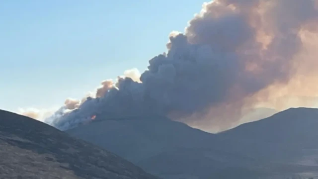 A wide shot of the huge plume of smoke coming from a wildfire at Sandbank Road, Hilltown, last Saturday. Three mountains are visible in the picture, one close up on the left and two in the distance, one of which is on fire. The smoking is spreading across a cloudless blue sky.