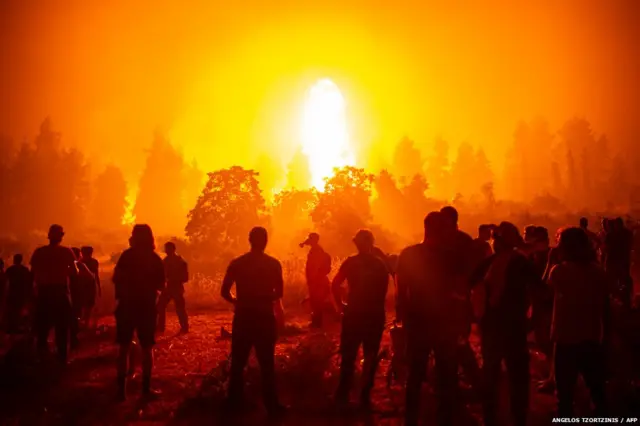 ग्रीस में लगे भयंकर आग को बुझाने में जुटे फ़ायरफ़ाइटर और उसे देखते स्थानीय लोग.