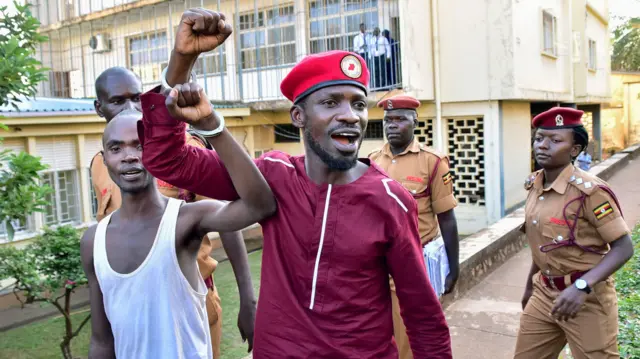 Bobi Wine lève le poing en l'air alors qu'il est menotté à un autre homme. Des personnes vêtues d'uniformes militaires marchent derrière eux.