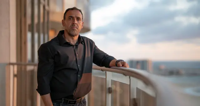 Eli Sharabi, vêtu d'une chemise noire, se tient sur un haut balcon d'un complexe d'appartements donnant sur une plage et la mer au coucher du soleil, son bras posé sur la balustrade du balcon tandis qu'il regarde la caméra.