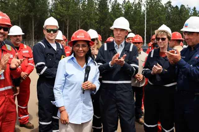 A presidente interina da Venezuela, Delcy Rodríguez, e o secretário da Energia dos Estados Unidos, Chris Wright, visitam instalações petrolíferas. Rodríguez usa capacete laranja e camisa azul clara e Wright usa um capacete branco e macacão azul