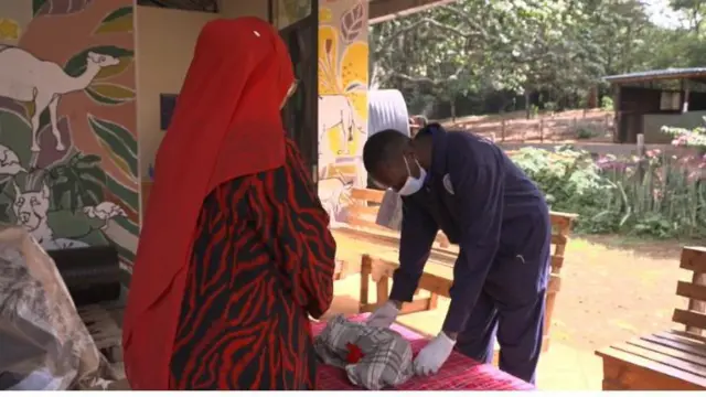 Mchoma maiti aliyevaa aproni ya rangi ya buluu akiwa amevaa glovu mikononi akiwa ameinama akitaka kumbeba huku mwanamke amevaa rinda jekundu akiangalia.