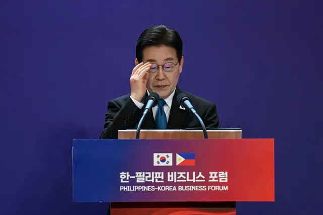 Presiden Korea Selatan Lee Jae Myung berada di atas podium yang memiliki simbol bendera negara Korea Selatan dan Filipina, sambil membenarkan letak kacamatanya, 