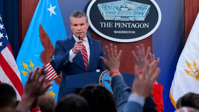 5 Mayıs’taki basın brifinginde mavi takım elbise ve kırmızı çizgili kravat giyen Hegseth, sağ elinde bir kalem tutarak işaret parmağıyla söz verdiği gazeteciyi gösterirken, muhabirlerin soru sormak için ellerini kaldırdığı görülüyor.