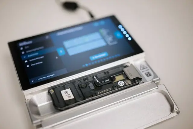 A imagem mostra um pequeno aparelho eletrônico de bancada, de cor metálica clara, com uma tela sensível ao toque inclinada na parte superior. A tela exibe uma interface escura com gráficos coloridos e menus laterais, sugerindo um software de análise. Logo abaixo dela, há um compartimento aberto onde está encaixado um cartucho retangular e fino, com etiquetas e pequenos componentes eletrônicos visíveis, incluindo conectores, parafusos e um chip preto. Esse cartucho ocupa quase todo o comprimento do aparelho e parece ser a área onde amostras são inseridas para sequenciamento. Um cabo preto está conectado à parte traseira, indicando que o equipamento está ligado.