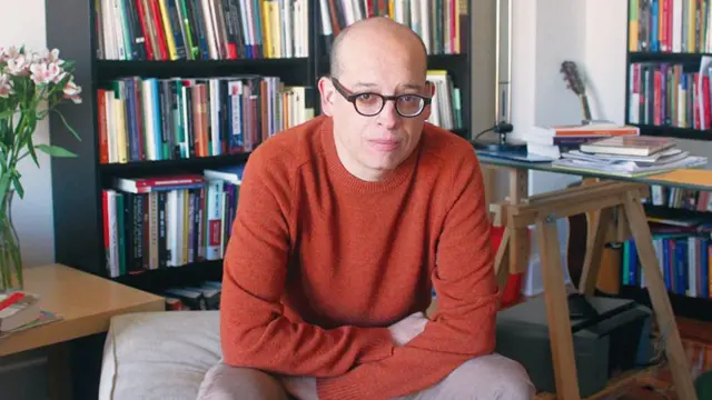 Alberto Vergara, sentado con brazos cruzados, con estantes con libros y una mesa de trabajo detrás.