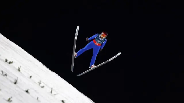 Muhammed Ali Bedir'in ayaklarında kayaklarla havada durduğu an, kamera aşağıdan çekiyor, geri plan karanlık. 