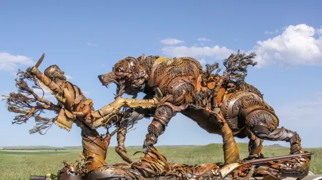Hibrit metalden yapılmış, bir bıçak tutan ve geri saldıran Hugh Glass'a saldıran bir boz ayıyı gösteren ünlü Hugh Glass anıtının bir fotoğrafı. Anıt kahverengidir, camdan ve ayıdan büyüyen ağaçların görünümü vardır. Parlak yeşil çimenli bir düzlemdedir. Gün güneşli, parlak mavi bir gökyüzü ve birkaç ince beyaz bulutla. 