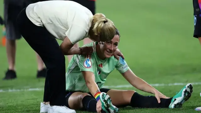 Sarina Wiegman agachada abraza a Lucy Bronze quien está sentada en la grama del campo de fútbol