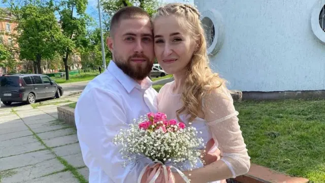 Katerina Malyshko (derecha) con su vestido de novia y sosteniendo un ramo de flores blancas y rosas, de pie junto a su esposo Vitaly, que lleva barba y una camisa blanca.