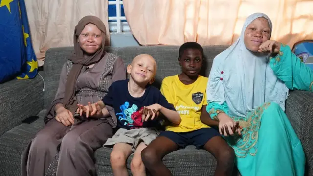 Three children with albinism and anoda without albinism sit in a sofa hand-in-hand and smiling to show their unity and love.