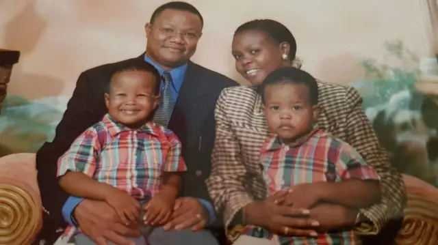 Mwanamume aliyevalia suti nyeusi, shati la samawati ameketi karibu na mwanamke aliyevalia shati la rangi la kahawia na nyeusi. Wanaangalia kamera huku wakitabasamu. Wamewapakata watototo wa kiume wawili waliovalia mashati yanayofanana.