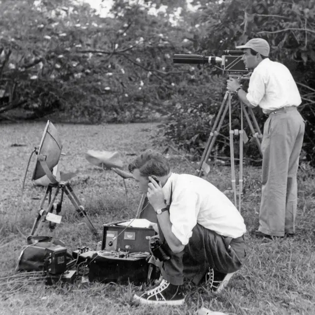 Fotografía en blanco y negro de dos personas grabando en exteriores, con una cámara en trípode y equipo de audio entre la vegetación.