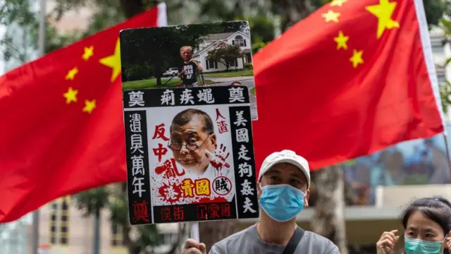 Manifestantes pro-Pekín sostienen un cartel que muestra a Jimmy Lai con una correa sostenida por Donald Trump en esta foto de 2021. Detrás de los manifestantes, ondean dos banderas chinas.