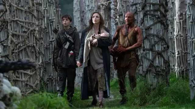 Tres personajes de la película "28 años después", una mujer cargando un bebé, un joven adolescente y un hombre de mediana edad. Caminan entre árboles adornados con fémures humanos.