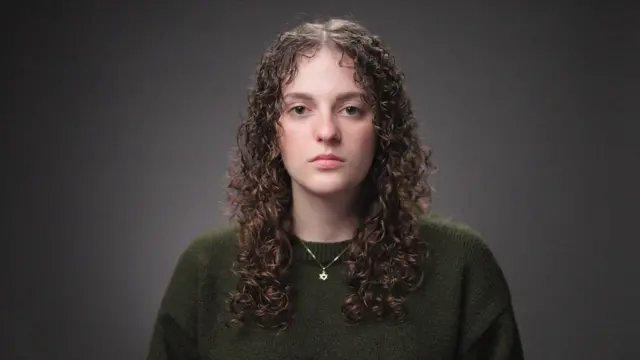 شابة تنظر إلى الكاميرا. لديها شعر طويل مجعد وترتدي سترة خضراء داكنة وسلسلة عليها نجمة داود حول رقبتها