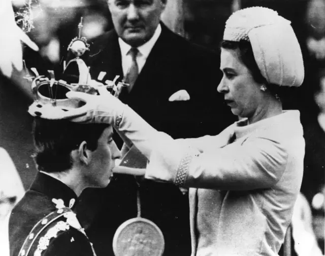 Queen Elizabeth II placing the coronet on Charles, Prince of Wales' head during his investiture ceremony whilst an official holds the Seal of Letters Patent. 