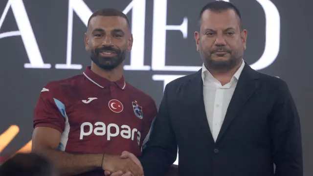 محمد صلاج يرتدي قميص نادي طرابزون التركي النبيذي وفي وسطه علم تركيا، يصافح رئيس نادي طرابزون سبور أرطغرل دوغان يرتدي سترة زرقاء داكنة وقميصاً أبيض اللون.