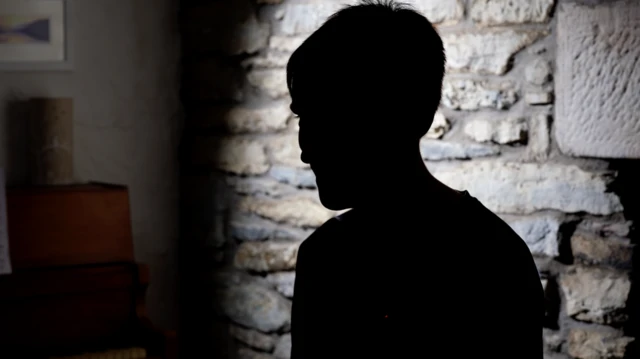 Silueta de un adolescente sentado de perfil contra un muro de piedra.