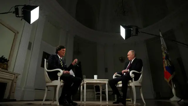 Карлсон и Путин сидят в белых креслах, рядом виден российский флаг с гербом, над ними софиты. 