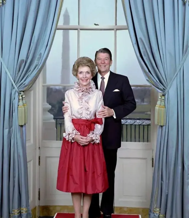 Nancy Reagan e Ronald Reagan posando para uma foto. Ela usa uma saia vemelha. Ele usa um terno. Ambos sorriem