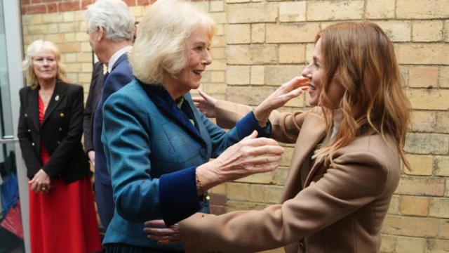 La reina Camila, con una elegante chaqueta de tweed azul y puños de terciopelo, extiende los brazos para saludar a Geri Halliwell, quien lleva una chaqueta de lana beige. Hay una pared de ladrillos detrás de ellas en un vestíbulo, donde esperan un par de dignatarios.