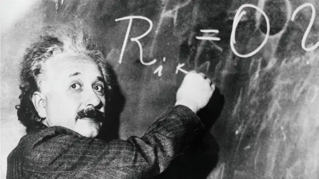 Albert Einstein mirando la cámara mientras escribe una fórmula matemática en un pizarrón.