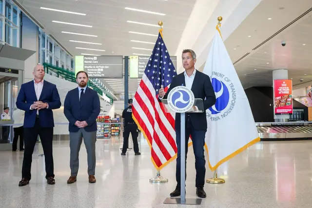 Министр транспорта США в синем деловой костюме делает заявление в аэропорту Ньюарка, на заднем плане видны табло вылетов и прилетов и багажные ленты