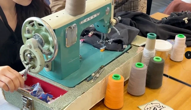 照片中是一位女子坐在老式縫紉機前，桌上擺滿彩色線軸。