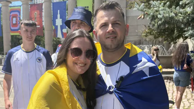 O casal Admir e Alisa Maric, usando bandeiras azuis e amarelas como capas, no lado externo de um estádio de futebol