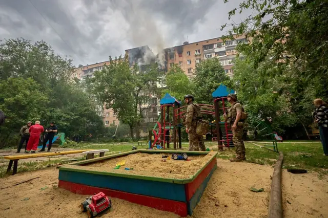 Vojnici i ljudi stoje na igralištu. Iza njih gori zgrada