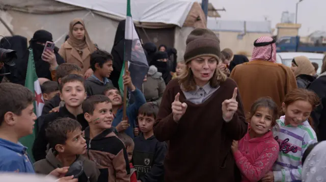 قبوات ترتدي سُترة بُنيّة اللون وقبعة صوفية، محاطة بنحو عشرة أطفال، ينظر إليها بعضهم. يبتسم بعضهم الآخر. تتحدث وتشير بسبّابتيها، وتظهر خلفها خيام نازحين.