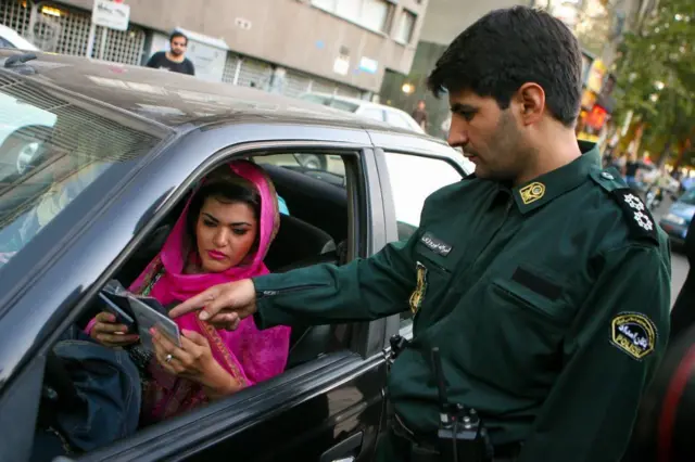 صورة لشرطة الأخلاق وهي توقف سيارة، تقودها سيدة تغطي رأسها بشال وردي اللون، ويظهر الجزء الأمامي من شعرها، بينما يقف ضابط يفحص أوراق بحوزة السيدة.