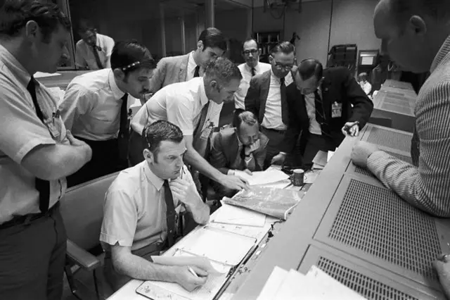 Controladores da missão Apollo 13 reunidos na sala de controle, estudando como fazer a espaçonave avariada e seus astronautas pousarem na Terra com segurança