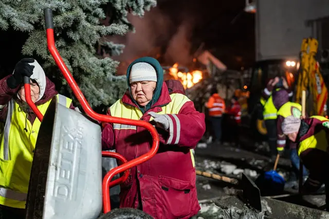 У центрі жінка середнього віку у червоній куртці тримає металеву тачку. Навколо видно інших працівників у світловідбивних жилетах, які прибирають уламки та працюють із лопатами. На задньому плані горять залишки будівлі, а поруч стоїть будівельна техніка, зокрема екскаватор.